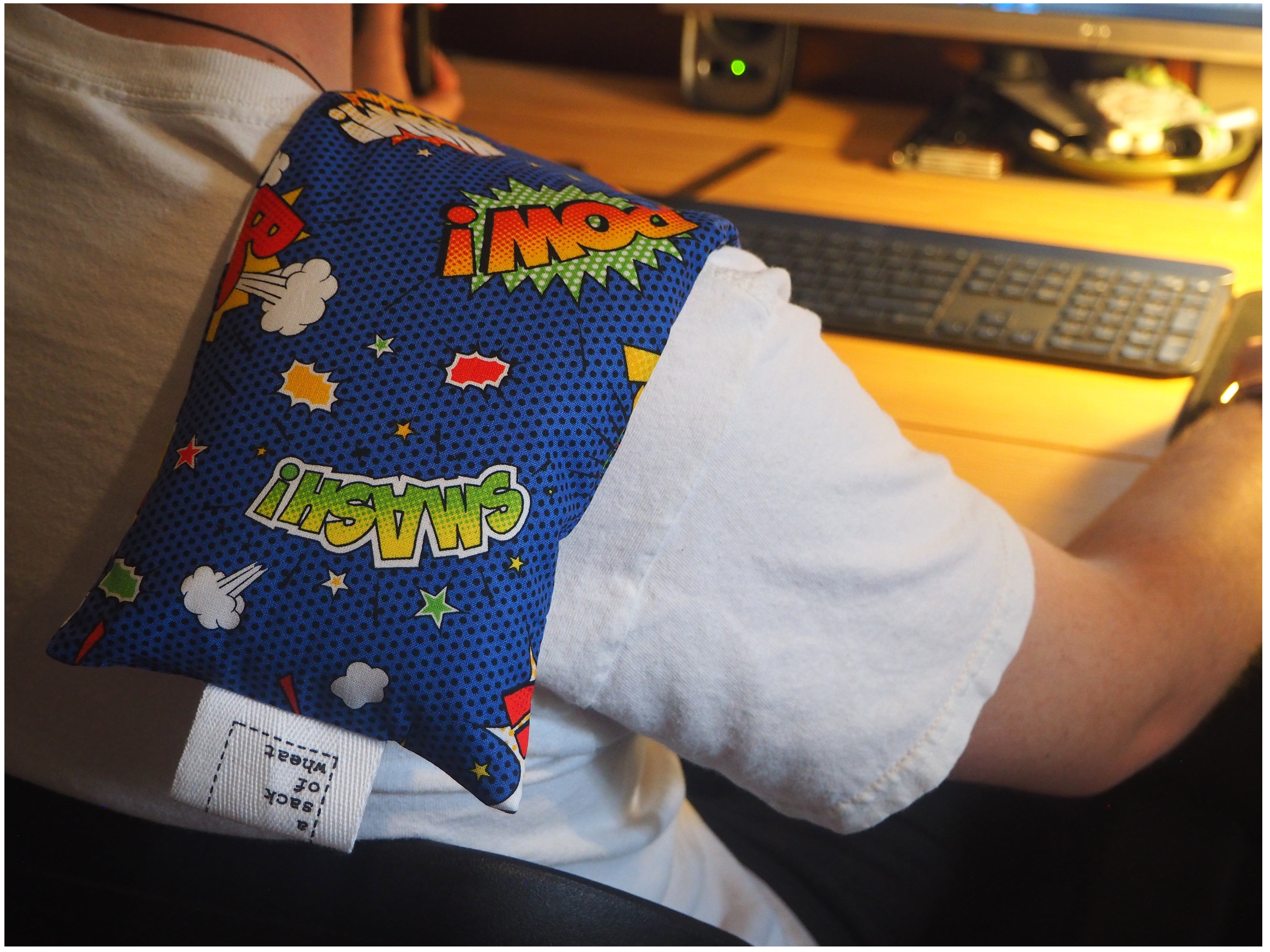 Colorful wheat bag with comic book print on mans shoulder in front of a computer setup.