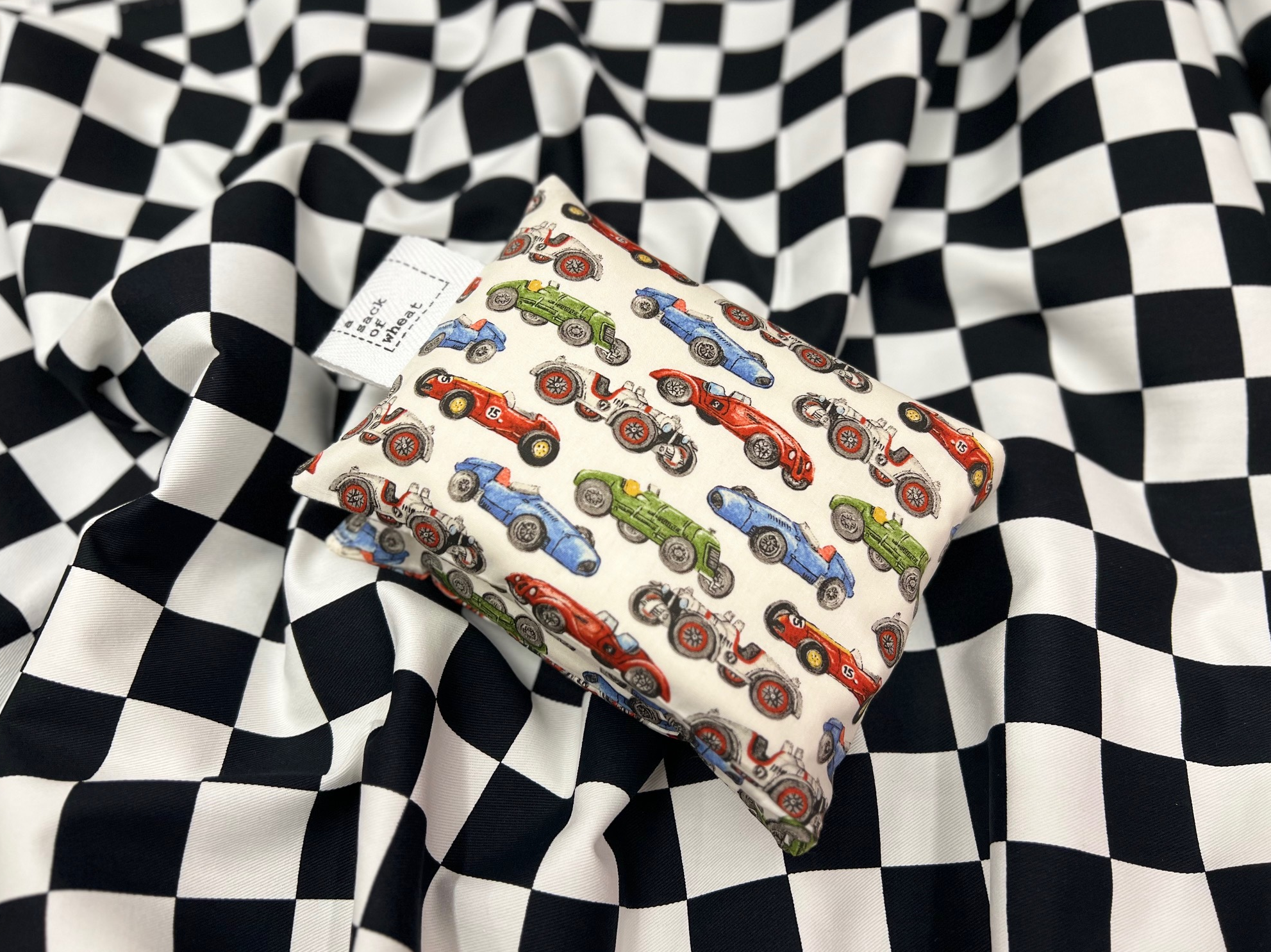 A folded wheat bag featuring images of colorful Vintage racing cars on cotton fabric. The wheat bag is photographed on a checkered fabric background.