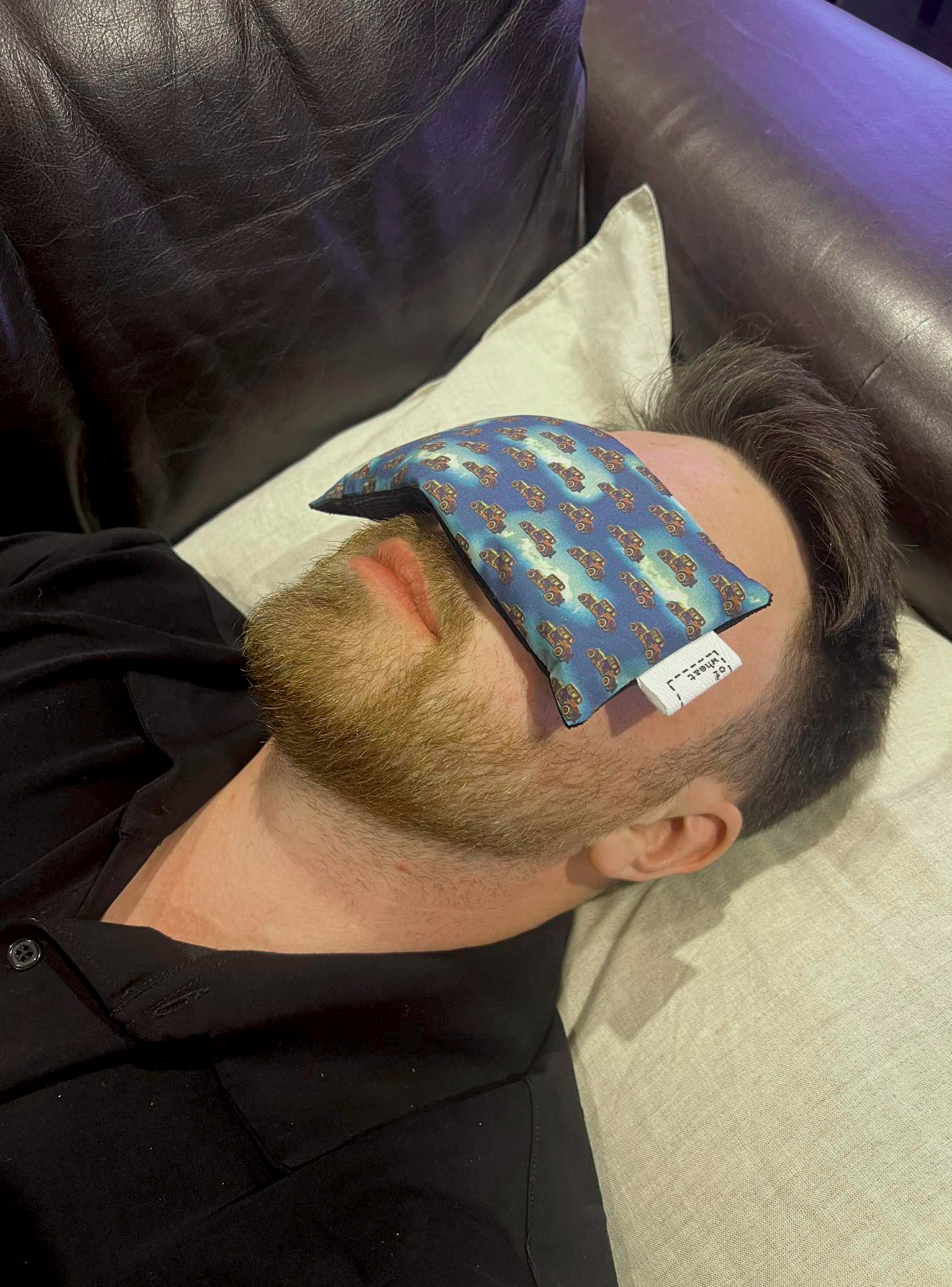 Person lying down with an eye pillow on their forehead, wearing a black shirt.